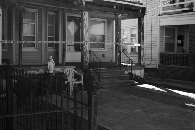 A black and white image of a suburban house with police tape obscuring its entrance.