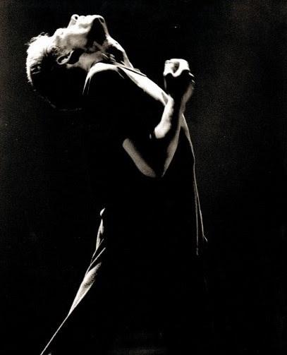 Black and white image of a young man mid 20's head thrown back looking at the sky face and chest lit by light from above his right arm bent with his hand in front if his chest.