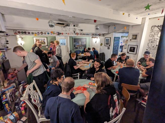 A room with people sitting a tables playing games. One person is looking at the selection of games and choosing one to play