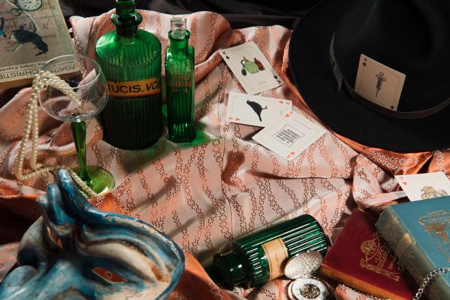 A satin table cloth covered in several mysterious looking objects. You can see a glass filled with pearls, a masquerade mask, poison bottles, playing cards and a fedora hat.