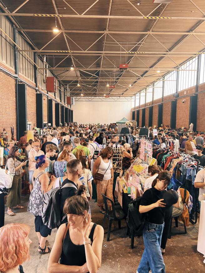 A crowd of people at an indoor flea market, shopping vintage clothing, furniture and craft.