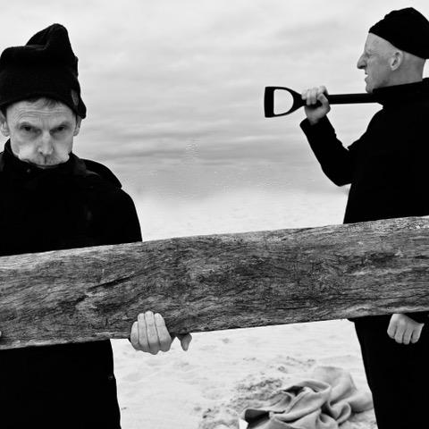 A black and white photograph of two performers wearing matching black clothing and hats. One carries a piece of wood and faces the camera, the other stands in profile, carrying a shovel over their shoulder.