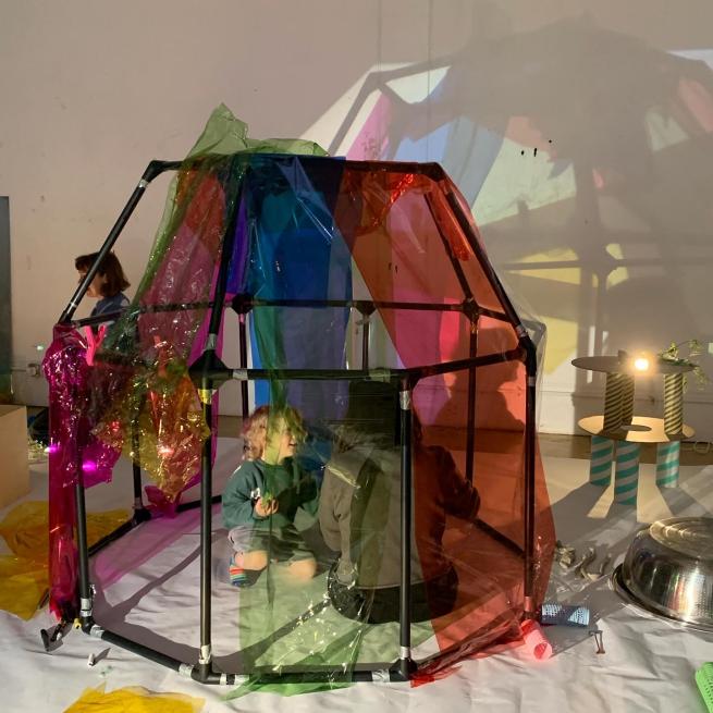 A young child and their parent are sat inside the frame of a den. Rainbow coloured cellophane is pegged to the den frame and the colours are projected onto the white wall, behind like stained glass. 