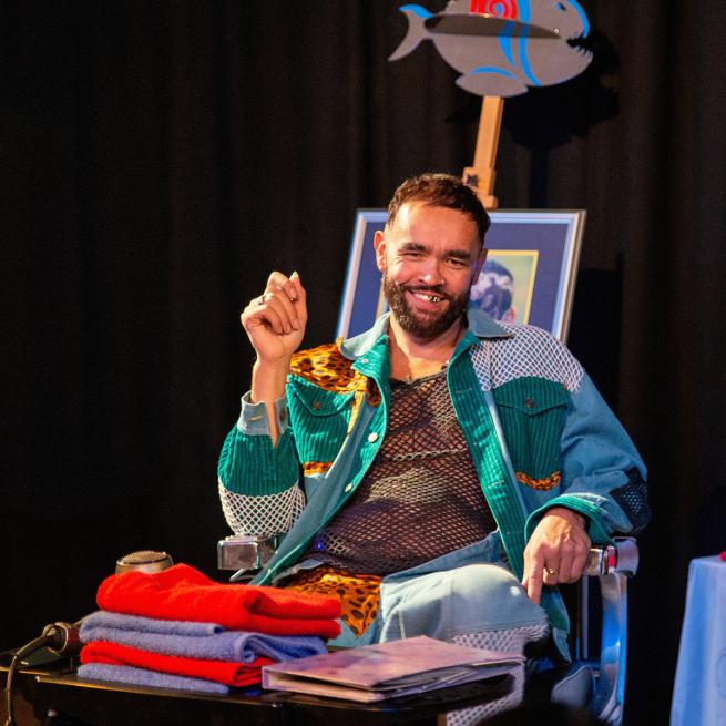  A man with a beard smiles while seated. He wears a colorful outfit and sits next to a stack of fabric. There's a framed picture and a fish decoration in the background.