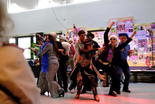 A group of people in colourful, 1970's themed costumes striking fun disco dance poses. They are wearing bold outfits, wigs, hats and sequins. The scene is lively, set against a background of retro posters, capturing the energy of a live performance, where the group of both men and women are fully immersed in the playful, celebratory moment.