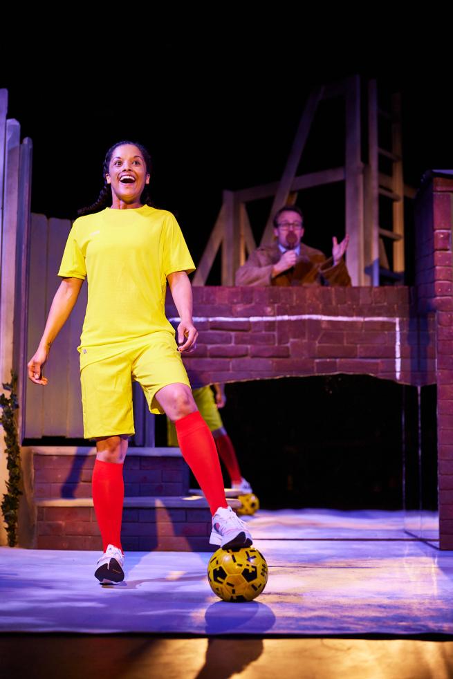 Freddie - played by Rhiannon Canoville-Ord, wearing bright yellow football kit, stand with one foot on yellow ball and a big smile on her face.