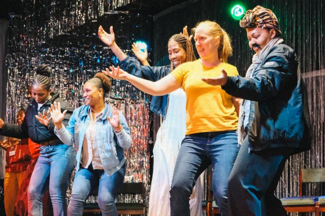 5 women dance together on a stage, they are facing towards an audience, there is a tinsel curtain behind them