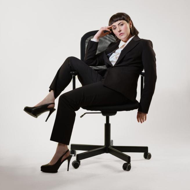 Emma Sidi sat casually sideways on an office chair wearing a black suit and black stilettos.