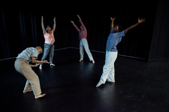 Four dances in a dance studio with a black dance floor and black out curtains behind them. Each dancer is striking a different pose and they are positioned in an open circle facing towards one another, with arms outstretched in different directions.