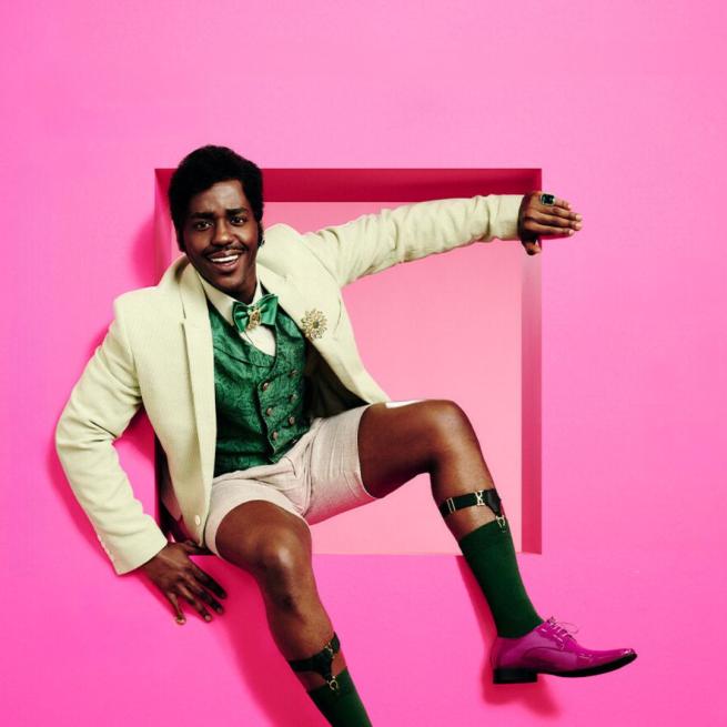 Ncuti Gatwa wearing a suit jacket, waist coat, bow tie and shorts sat in a cut out in a pink wall.