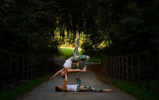 2 ladies, dressed for and performing acroyoga, one is holding the other up with her feet, whilst the on on top strikes a pose with a far back reaching stretch. Setting is in a park on a path, shaded under trees.