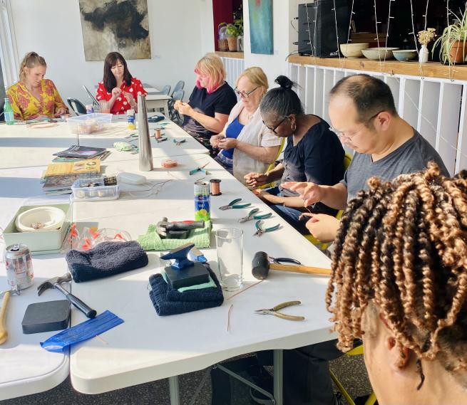 A diverse group of people sat concentrating around a table with tools making jewellery. 