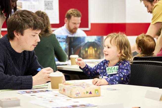 Children and her parent/carer playing a board game with other families playing around them.