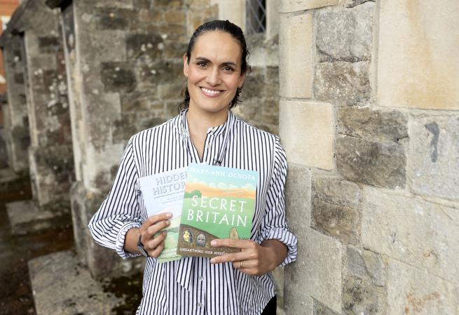 Author Mary Ann Ochota holding two of her books by a stone wall