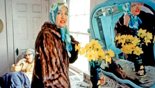 Edie in a blue headscarf and big fur coat looking in a mirror with her reflection in the mirror and some yellow flowers by the mirror with Edith sitting behind