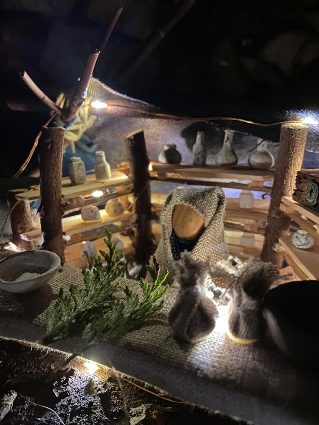 Photo of a model of a tiny shopkeeper standing behind the counter of her shop. There are wooden shelves in the background with bottles on them. On the counter there are goods for sale.