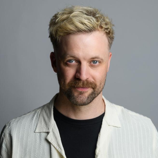 Headshot of Thomas Green wearing a black t-shirt under a pale cream light shirt stood against a grey background.