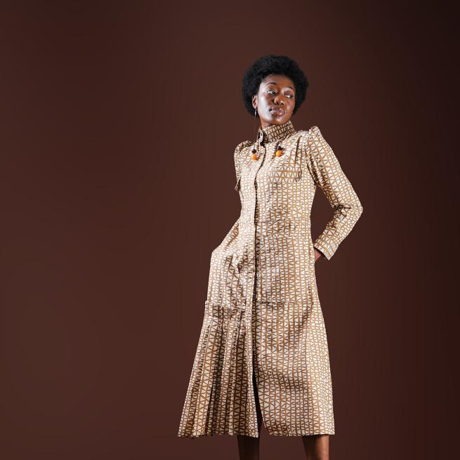 A black woman poses wearing cream patterned dress in from of brown background 