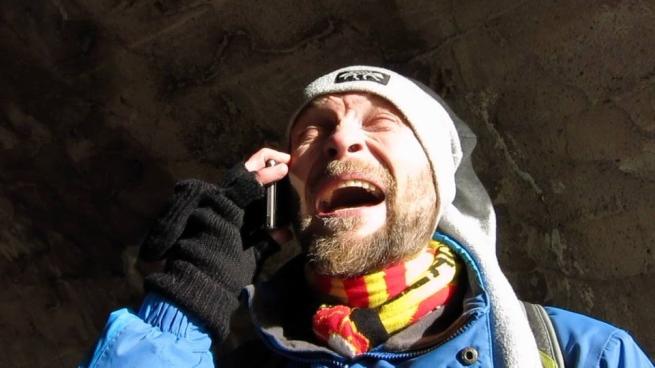 man wrapped up in many layers, coat, hat, scarf. His face appears to be laughing whilst on a phone. His face has a warm light on it against a dark backfrop