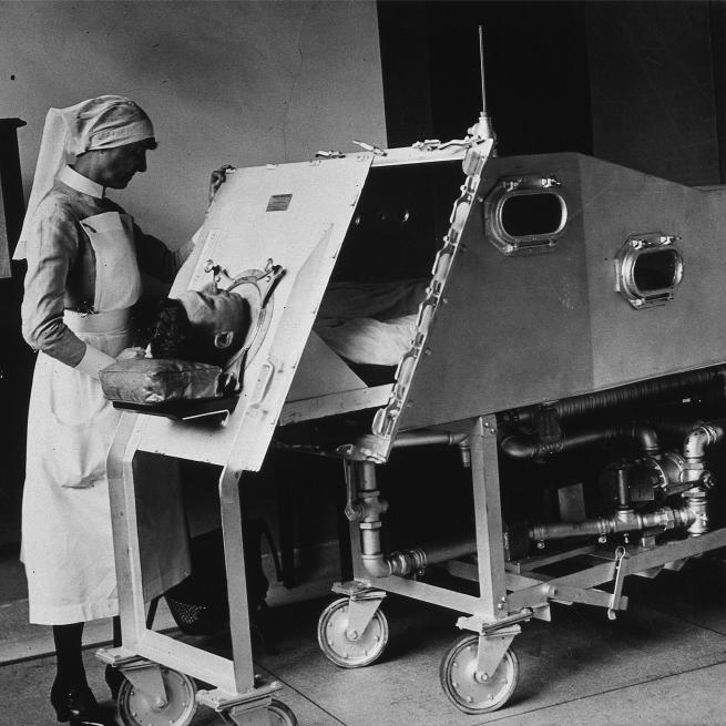  A black and white image of a nurse stood beside a man inside an iron lung.