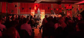 A person with a microphone stands on a stage in a red lit room, performing to a large crowd.