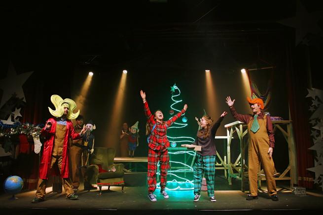 Picture of Codswallop Christmas performance. Four people are stood in front of a Christmas scene with a light up Christmas tree, elf puppets and fireplace. The actors have their hands in the air and have happy faces.