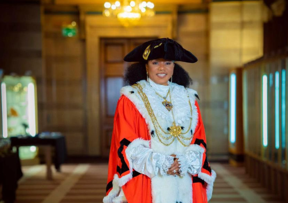 The Lord Mayor Abigail Marshall Katung, a black woman of Nigerian heritage, stands smiling in Civic Hall in Leeds. She is wearing in her ceremonial Lord Mayor's robes in red and black with a white fur collar, her gold mayoral chain and a black tri-corn hat