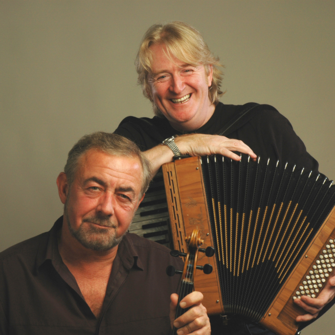 One man smiling whilst holding an accordion whilst the other is holding the top of a fiddle.