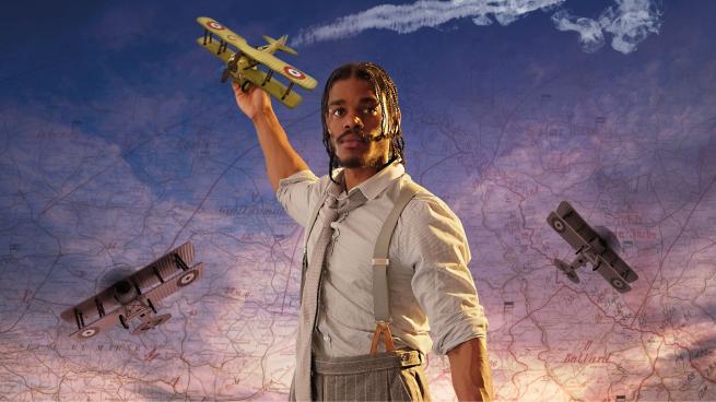 A person in a shirt, tie and dungarees holding a prop plane. The background is a blue and purple sky.