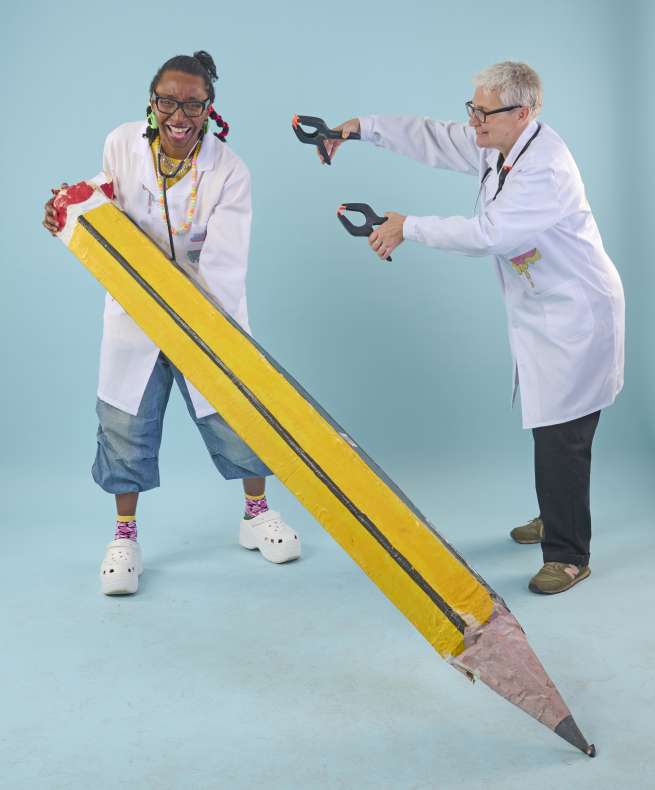 A smiling woman wearing a white lab coat holds a giant yellow pencil. To her right, another woman wearing a white lab coat reaches out playfully with a pair of giant pliers.