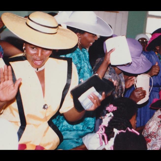 Women in a selection of church hats raise their hand holding their purses 