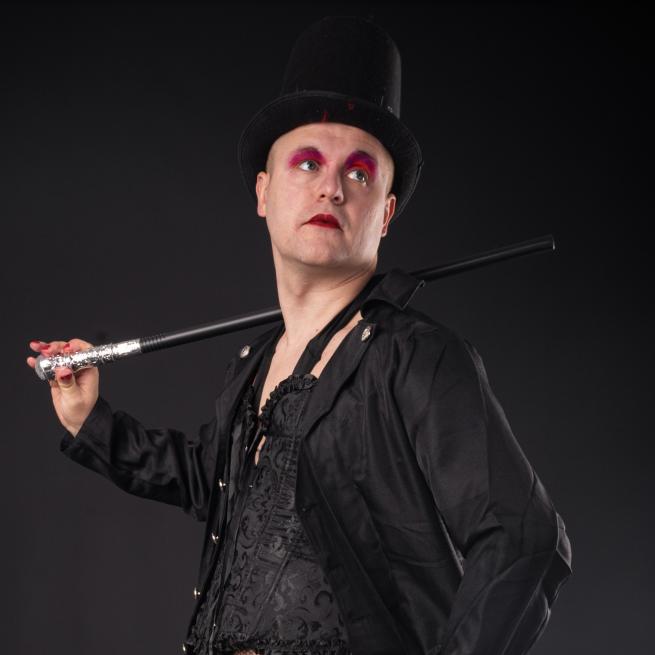 A man looking up with a black top hat, a black and sliver cane resting on his shoulder, pink eyeshadow and lipstick and a black top and jacket