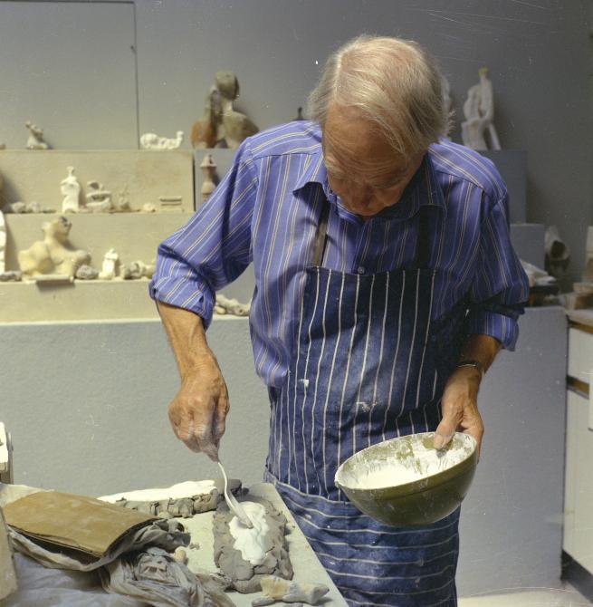 Henry Moore wears a stripy blue shirt and stripy blue apron as he mixes plaster and pours it into a mould.