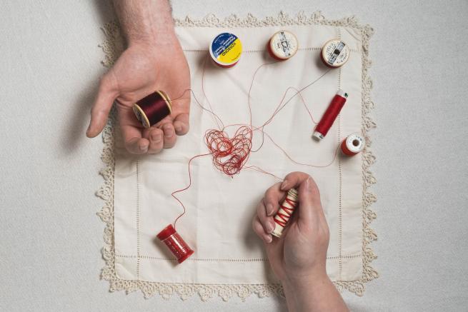 Two hands hold threads resting on an embroidered heart on a square of fabric