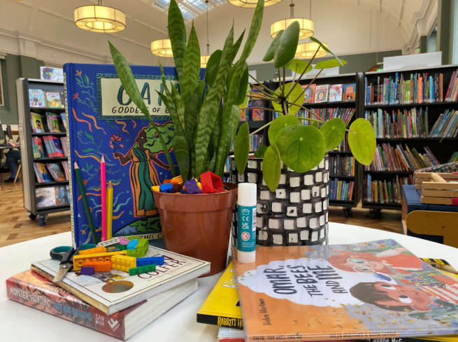 Plants, children's books and craft materials on a table in a library.