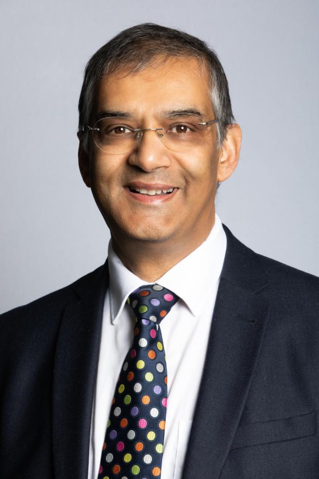 A headshot of a middle aged south Asian man smiling wearing a suit and spotty tie with glasses on