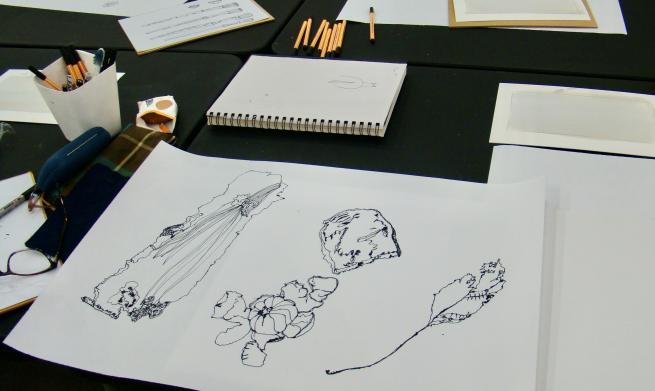 A table with paper,  pencils and an open sketchbook showing  pen drawings