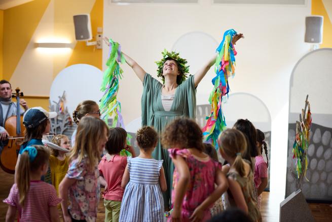 Children gathered round the mountain witch, her arms are in the air with colourful ribbons hanging down from her hands.