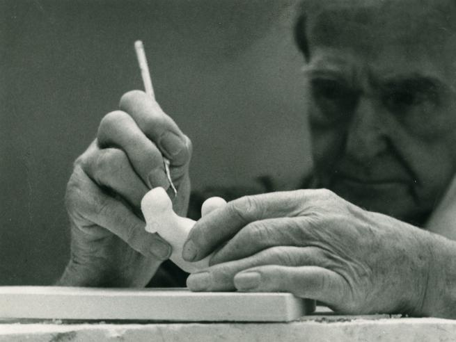 Black and white photograph with Henry Moore holding a plaster maquetter and working with a tool on it.