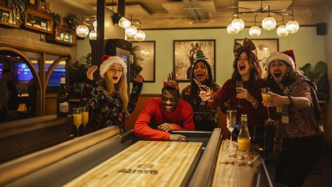 A group of people playing shuffleboard at Electric Shuffle, while wearing Christmas hats and jumpers