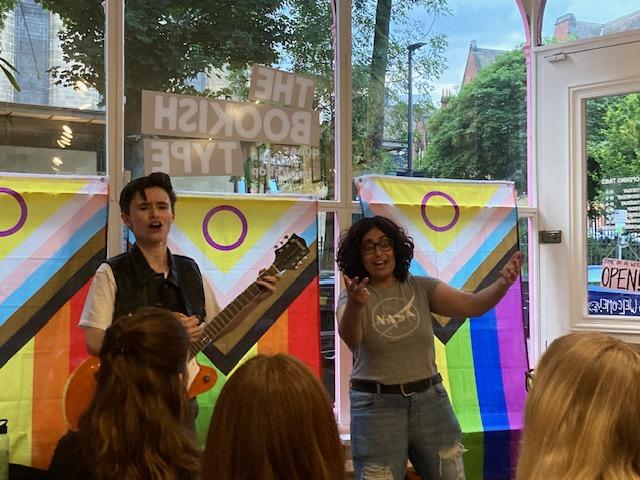 Two people stand in front of Pride flags performing a song