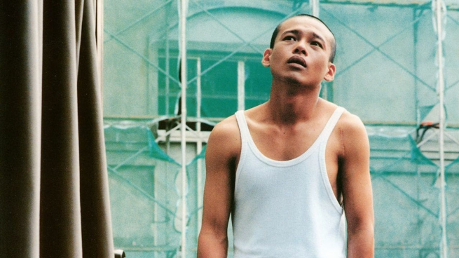 A man stands in the middle of shot infront of scaffolding wearing a white vest