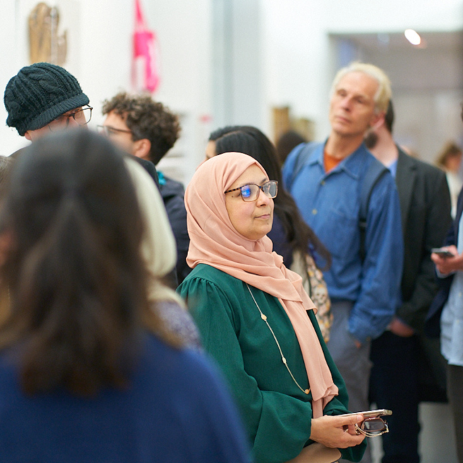 A woman wearing glasses and a headscarf looks at a work of art on a wall, surrounded by other gallery-goers