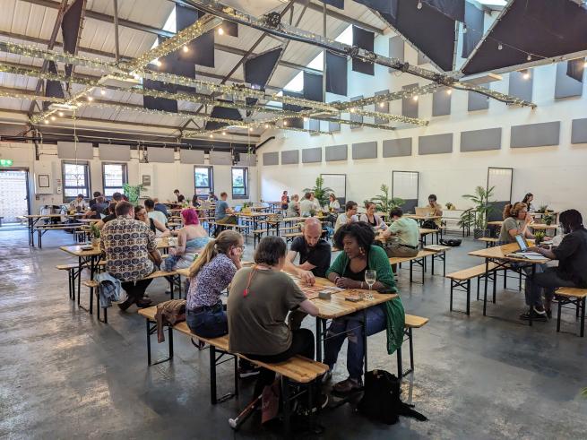 A large room with a high ceiling and fairy lights high up. People are sitting on benches at tables and playing boardgames. 