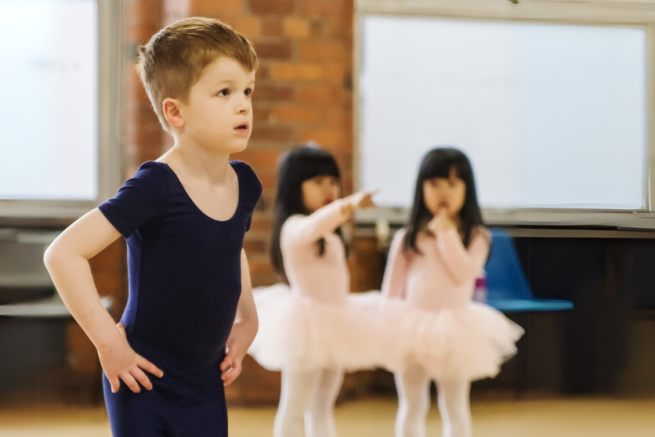 A little boy in the foreground in a navy blue leotard, in first position with two little girls in pink tutus out of focus behind him