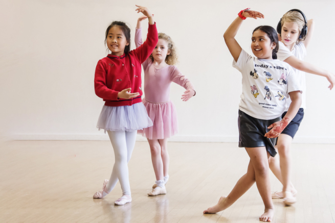 four girls dancing in a ballet position with one arm over their head and the other across them. Two girls in tutus and two in shorts and t-shirts, two girls stand at the front with the girl in shorts on the left smiling happily