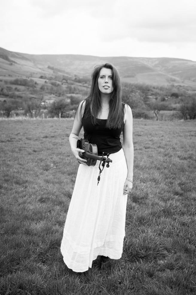 Black and white image of a woman wearing a white skirt and black top with long brown hair stood in a field holding a fiddle.