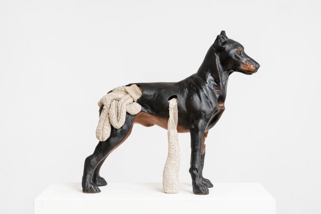 A sculpture of a dark brown dog looks to the right, hanging out of its middle and the back are what appears to be its insides (in cream) spilling out.