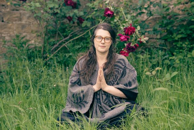 sophie sat on the grass in front of a tree and bush. She's looking at the camera, with her hands together, sat crossed legged, inviting you to join yoga