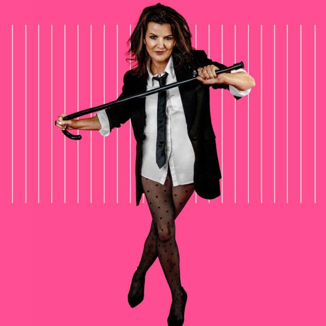 Deirdre O'Kane in a suit jacket, black tie, white shirt and just semi-sheer tights and black heels holding a cane across her chest with both hands stood against a pink background.